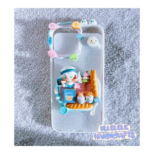 May include: A white phone case with a 3D character design. The character is a small person wearing a blue and white sailor outfit and holding a book that says "Snooks". The case also has a variety of other 3D elements, including a baguette, a cloud, and a small bear.