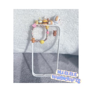 May include: A white phone case with a brown and white patterned design. The case has a variety of small charms attached to it, including a bear, a hamburger, a drink, and a dog. The text "BLUEUE HANDICRAFTS" is visible in the bottom right corner of the image.