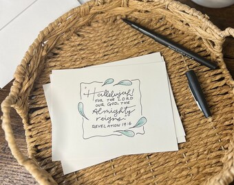 Op de afbeelding: Een natuurlijke vlechtmand bevat witte kaarten met de tekst "Hallelujah! For the Lord our God, the Almighty reigns." Een zwarte pen ligt op de kaarten. Witte enveloppen en een plant in een pot staan op de achtergrond.