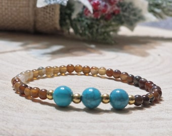 May include: A beaded bracelet featuring alternating brown and amber-colored beads with three turquoise-colored beads and gold-colored accents. The bracelet is displayed on a wooden surface, with a blurred background of decorative elements.