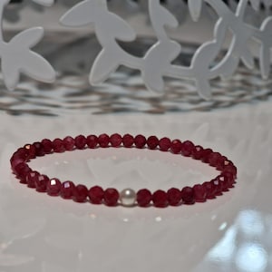 Peut inclure: Un bracelet délicat composé de petites perles rouge foncé à facettes. Le bracelet comporte une seule perle blanche, ronde. Les perles sont enfilées pour former un bracelet souple. Le bracelet est présenté sur une surface blanche.