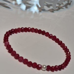 May include: A beaded bracelet featuring small, faceted, deep red beads. The bracelet includes a single, round, white pearl. The beads are strung together to form a flexible band, suitable for wearing on the wrist. The bracelet is displayed on a white surface.