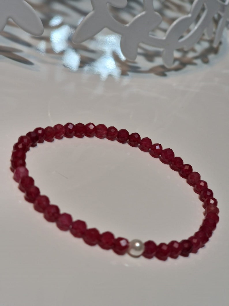 May include: A beaded bracelet featuring small, faceted, deep red beads. The bracelet includes a single, round, white pearl. The beads are strung together to form a flexible band, suitable for wearing on the wrist. The bracelet is displayed on a white surface.