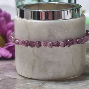 May include: A purple amethyst gemstone bracelet with small round beads. The bracelet is resting on a white marble surface.