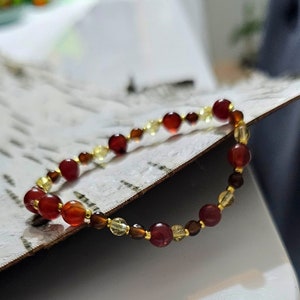 Könnte beinhalten: Ein Armband mit roten, bernsteinfarbenen und gelben Perlen in verschiedenen Größen. Die Perlen sind auf goldfarbenem Metall aufgefädelt. Das Armband wird auf einer strukturierten Oberfläche präsentiert.