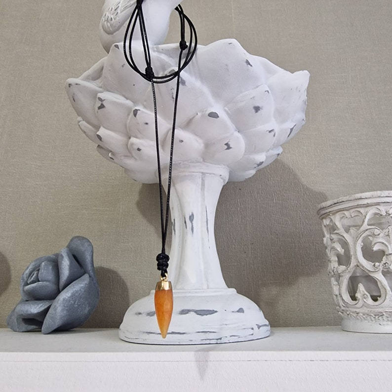 May include: A long, orange, pointed pendant hangs from a black cord necklace. The pendant is attached to a gold-colored connector. The necklace is hanging from a white ceramic bird statue.