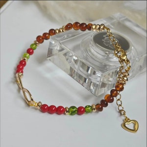 Könnte beinhalten: Ein goldfarbenes Armband mit einem Unendlichkeits-Symbol-Anhänger und roten, grünen und braunen Perlen. Das Armband hat einen herzförmigen Anhänger am Ende.