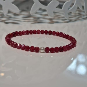 May include: A delicate bracelet featuring small, faceted, deep red beads. A single, round, white pearl is centered in the design. The bracelet is displayed on a white surface, with a decorative white metal accent in the background.