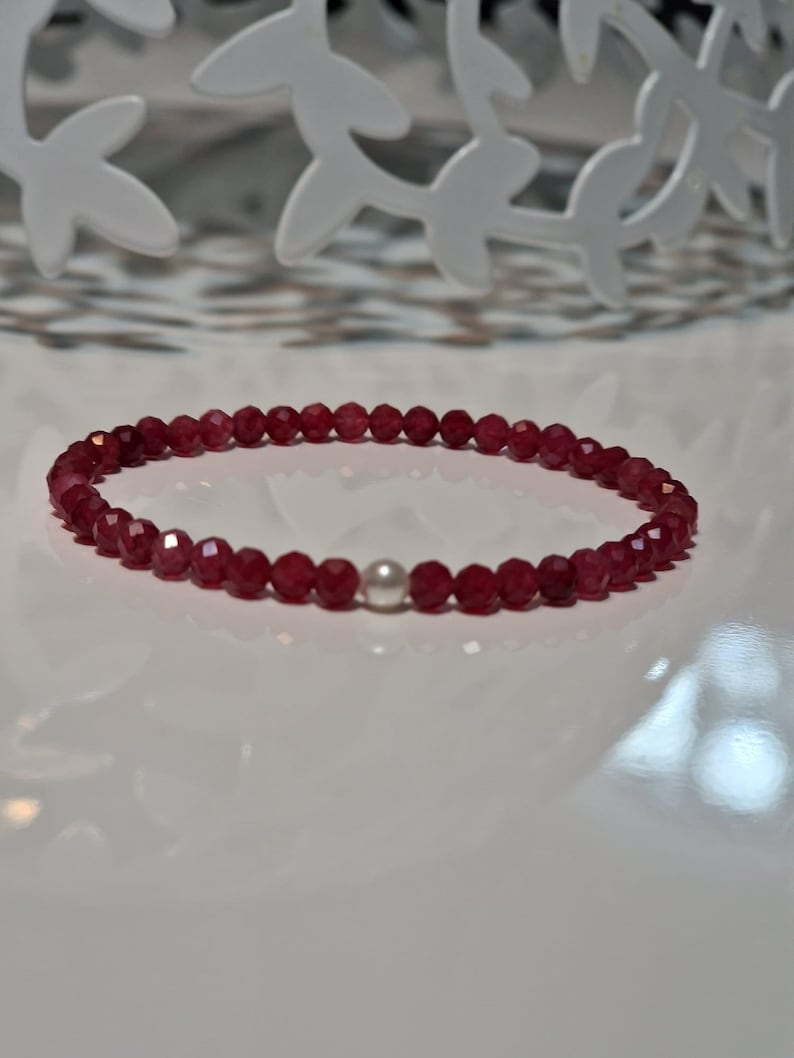 May include: A delicate bracelet featuring small, faceted, deep red beads. A single, round, white pearl is centered in the design. The bracelet is displayed on a white surface, with a decorative white metal accent in the background.