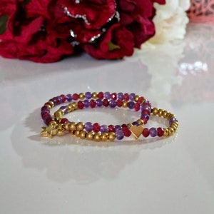 Könnte beinhalten: Zwei goldene Perlenarmbänder mit violetten und roten Edelsteinperlen. Ein Armband hat einen goldenen Sternanhänger und das andere einen goldenen Herzanhänger.