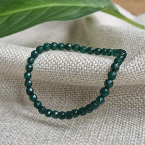 Pode incluir: Um bracelete de pedras preciosas verdes feito de contas facetadas. O bracelete está sobre um fundo de tecido castanho claro.