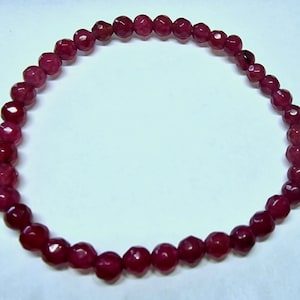 May include: A deep red beaded bracelet. The beads are faceted, giving them a sparkling appearance. The bracelet is strung together, forming a complete circle. The beads are a rich, dark red color.