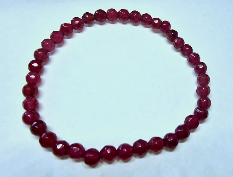 May include: A deep red beaded bracelet. The beads are faceted, giving them a sparkling appearance. The bracelet is strung together, forming a complete circle. The beads are a rich, dark red color.