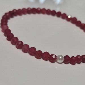 May include: A delicate bracelet featuring faceted, deep red beads. A single, round, white pearl bead is interspersed among the red beads. The bracelet is strung on a thin, flexible cord, suitable for everyday wear.