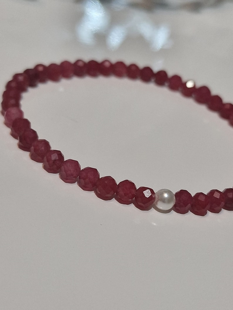 May include: A delicate bracelet featuring faceted, deep red beads. A single, round, white pearl bead is interspersed among the red beads. The bracelet is strung on a thin, flexible cord, suitable for everyday wear.