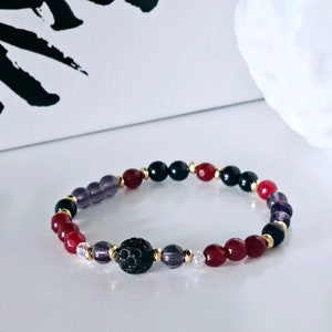 Könnte beinhalten: Ein Armband mit roten, schwarzen, violetten und durchsichtigen Perlen. Das Armband hat eine schwarze Lavaperle in der Mitte.