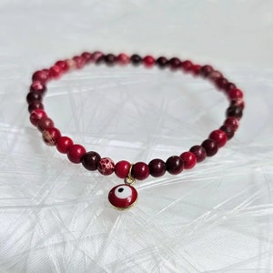 Può includere: Un braccialetto di perline rosse e bordeaux con un ciondolo a forma di occhio turco dorato.