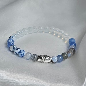 Peut inclure: Un bracelet en agate bleu et blanc avec un charme en argent. Le bracelet est composé de petites perles rondes et présente un dégradé de couleurs, passant du blanc au bleu.