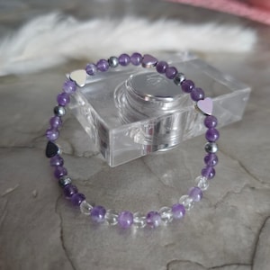Könnte beinhalten: Ein violettes und klares Kristallperlenarmband mit silbernen Herzanhängern. Das Armband liegt auf einer weißen Oberfläche.