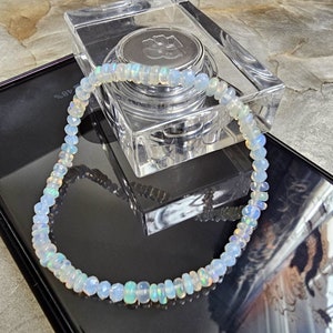 Peut inclure: Un collier délicat composé de perles d'opale iridescentes. Les perles présentent un mélange de blanc, de bleu et d'autres couleurs. Le collier est présenté sur une surface noire, avec un récipient carré transparent en arrière-plan.