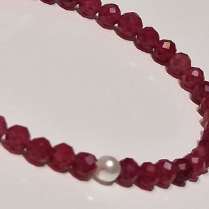 May include: A necklace featuring faceted, deep red beads with a single, round, white pearl. The beads are strung together, creating a delicate and elegant piece of jewelry. The necklace is displayed against a white background.