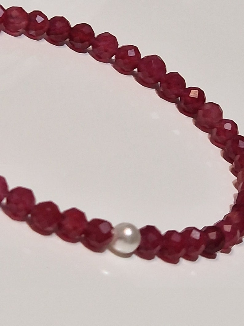 May include: A necklace featuring faceted, deep red beads with a single, round, white pearl. The beads are strung together, creating a delicate and elegant piece of jewelry. The necklace is displayed against a white background.