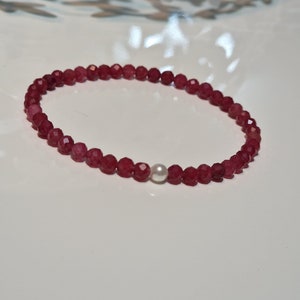 May include: A beaded bracelet featuring faceted, deep red gemstones and a single, round, white pearl. The bracelet is strung on an elastic band and is displayed on a white surface.