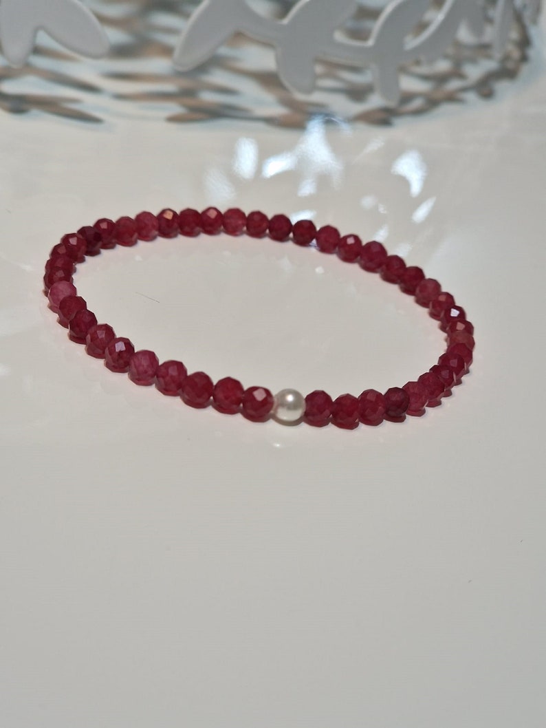 May include: A beaded bracelet featuring faceted, deep red gemstones and a single, round, white pearl. The bracelet is strung on an elastic band and is displayed on a white surface.