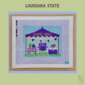 May include: A needlepoint canvas featuring a purple tent with the text GAME DAY, yellow and purple bunting, a table with a checkered cloth, a purple chair labeled LSU, and a cooler with a football on top, all set against a light blue background.