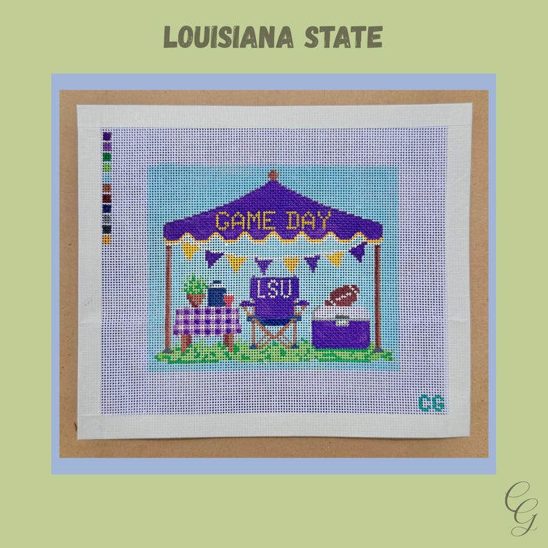 May include: A needlepoint canvas featuring a purple tent with the text GAME DAY, yellow and purple bunting, a table with a checkered cloth, a purple chair labeled LSU, and a cooler with a football on top, all set against a light blue background.