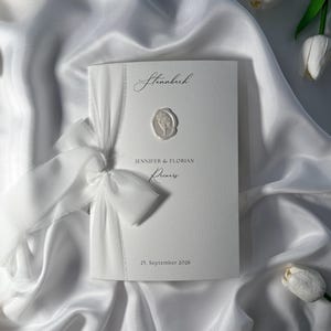 Può includere: Un invito di nozze bianco con un nastro e un fiocco, con i nomi "Jennifer & Florian" e la data "25. September 2026". L'invito presenta un sigillo di cera floreale e il nome "Stannbach" in alto.