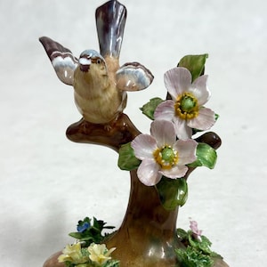 May include: A detailed porcelain figurine of a bird perched on a tree branch with pink flowers and green leaves. The bird has brown, white, and blue feathers. The base features yellow and blue flowers.