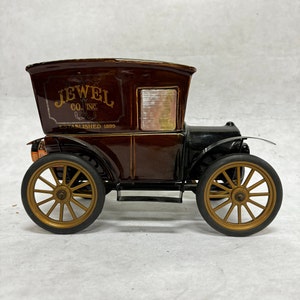 Puede incluir: Un coche de juguete vintage marr&oacute;n y negro con ruedas doradas. El coche tiene el texto "JEWEL CO. INC. ESTABLISHED 1899" en el lateral. El coche tiene un dise&ntilde;o cl&aacute;sico, con una zona de pasajeros cubierta y un cap&oacute; delantero.