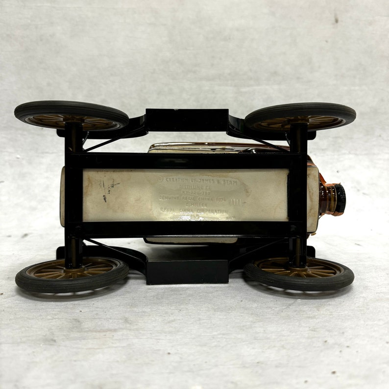 Puede incluir: Una botella de licor de estilo vintage con forma de coche cl&aacute;sico. El marco negro sostiene la botella, con cuatro ruedas y un tap&oacute;n marr&oacute;n. La botella tiene el texto "Creation of James B. Beam".