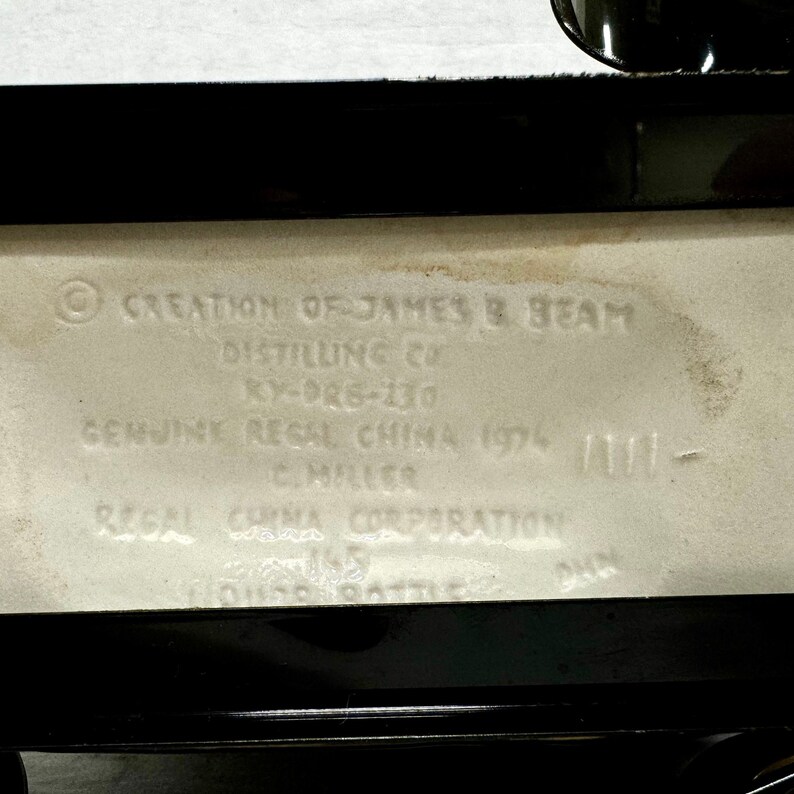 Puede incluir: Primer plano de la base rectangular de una botella de licor de cer&aacute;mica blanquecina con ribete negro. El texto incluye "CREATION OF JAMES B BEAM DISTILLING CO" y "GENUINE REGAL CHINA 1974".