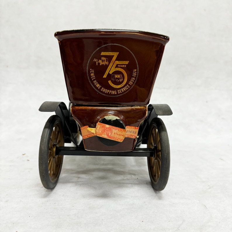 Puede incluir: Un recipiente de cer&aacute;mica marr&oacute;n con forma de coche antiguo con ruedas doradas. El recipiente tiene un logotipo que dice "Jewel Home Shopping Service 75 Years 1899-1974". El coche tiene guardabarros negros y una pegatina en la parte delantera.