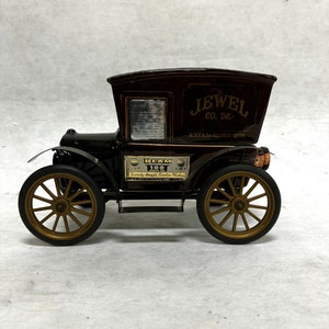 Puede incluir: Un coche de juguete de estilo vintage con la parte superior marr&oacute;n y el cuerpo negro. El coche tiene ruedas doradas y el texto "JEWEL CO. INC. ESTABLISHED 1899" en el lateral. Tambi&eacute;n tiene una etiqueta "BEAM 100 MONTHS".