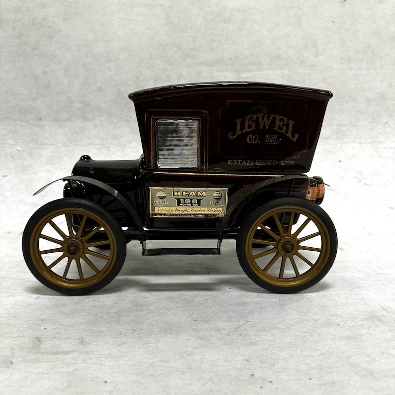 Puede incluir: Un coche de juguete de estilo vintage con la parte superior marr&oacute;n y el cuerpo negro. El coche tiene ruedas doradas y el texto "JEWEL CO. INC. ESTABLISHED 1899" en el lateral. Tambi&eacute;n tiene una etiqueta "BEAM 100 MONTHS".
