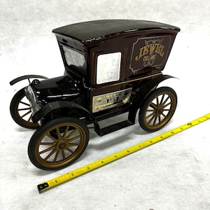 Puede incluir: Una licorera vintage en forma de coche negro y marr&oacute;n con detalles dorados. La licorera tiene un techo texturizado y presenta el texto "JEWEL CO. INC. ESTABLISHED 1898" y "BEAM 100". El coche tiene ruedas de radios doradas.
