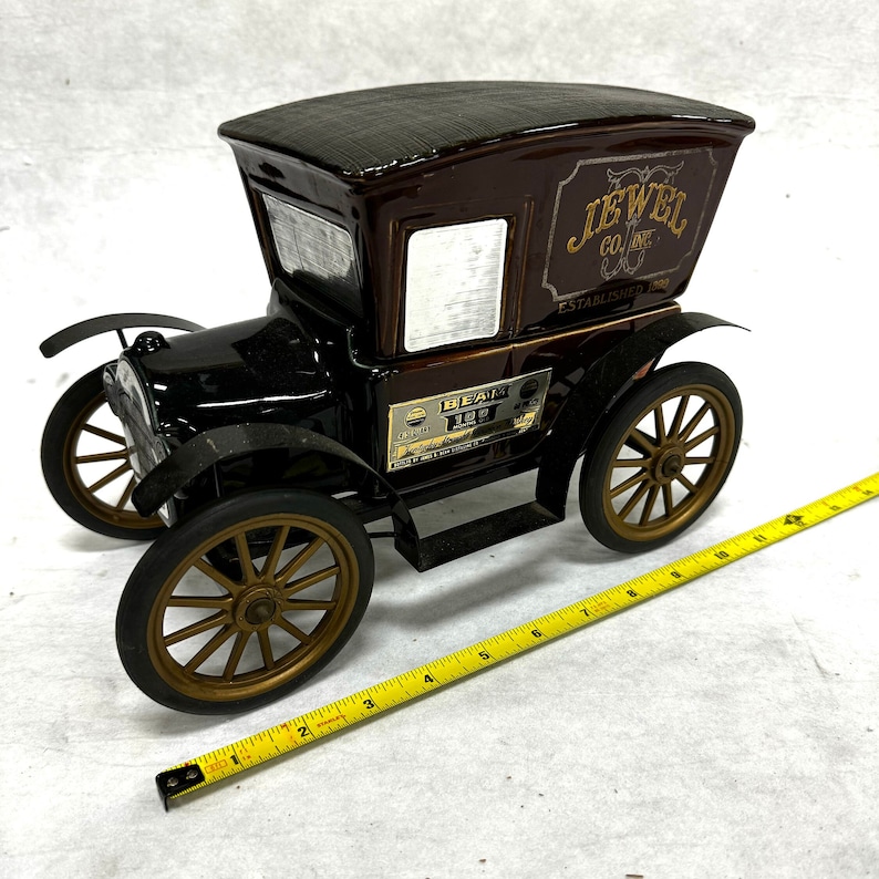 Puede incluir: Una licorera vintage en forma de coche negro y marr&oacute;n con detalles dorados. La licorera tiene un techo texturizado y presenta el texto "JEWEL CO. INC. ESTABLISHED 1898" y "BEAM 100". El coche tiene ruedas de radios doradas.