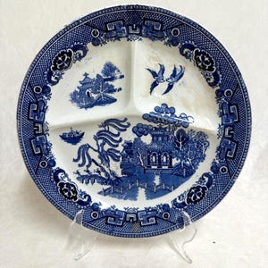 May include: A blue and white divided plate with a classic Willow pattern. The plate features a central design of a pagoda, bridge, and trees, with additional scenes and decorative borders. The plate is supported by a clear plastic stand.