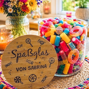 Op de afbeelding: Een glazen pot vol kleurrijke gummy snoepjes en een houten deksel met de tekst "Spaßglas von Sabrina" en snoep illustraties. De pot staat op een geweven mat met een kleurrijke pompom rand, met een vaas met bloemen op de achtergrond.