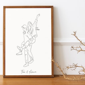 May include: A black and white line drawing of a couple, a woman with her arms raised and a man carrying her on his back. The drawing is on a white background and framed in a brown wooden frame. The text "Tom & Anna" is written below the drawing.