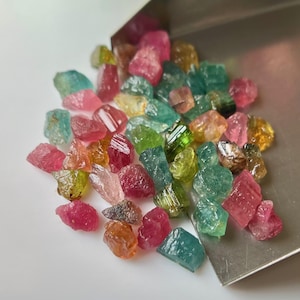 May include: A collection of colourful, raw tourmaline gemstones in various shapes and sizes. The gemstones are a mix of pink, green, and blue.