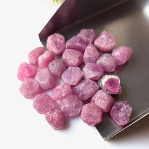 May include: A collection of rough, pink ruby gemstones. The gemstones vary in shade from light to deep pink, with a few showing a mix of pink and white. They are displayed on a silver tray against a white background.
