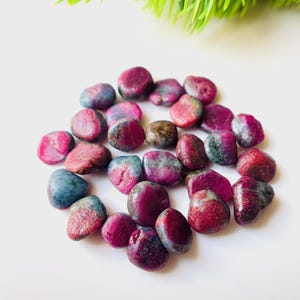 Op de afbeelding: Een verzameling gepolijste robijn edelstenen in tinten rood, roze en blauw. De edelstenen zijn glad en afgerond, met een natuurlijke, organische vorm. De stenen zijn verspreid op een wit oppervlak, met een vleugje groen gebladerte op de achtergrond.