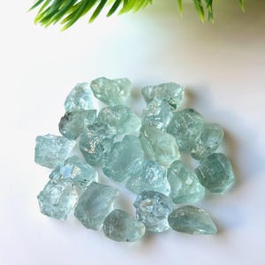 May include: A collection of light blue aquamarine rough stones. The stones are various sizes and shapes, with some having a more translucent appearance than others.