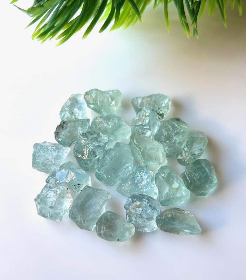 May include: A collection of light blue aquamarine rough stones. The stones are various sizes and shapes, with some having a more translucent appearance than others.