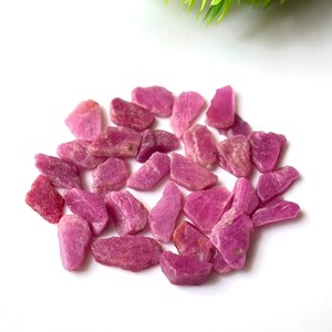 Könnte beinhalten: Eine Ansammlung kleiner, roher, unpolierter Rubin-Edelsteine in verschiedenen Schattierungen von leuchtendem Pink und tiefem Magenta. Diese natürlichen Mineralfragmente weisen unregelmäßige, gezackte Formen und texturierte Oberflächen auf.