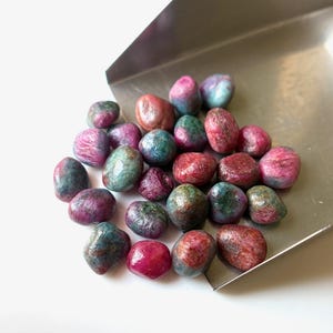 Può includere: Una collezione di pietre preziose lucide nei toni del rosso, rosa e verde acqua, presentate su un vassoio d'argento. Le pietre hanno una forma liscia e arrotondata e una lucentezza leggermente iridescente.